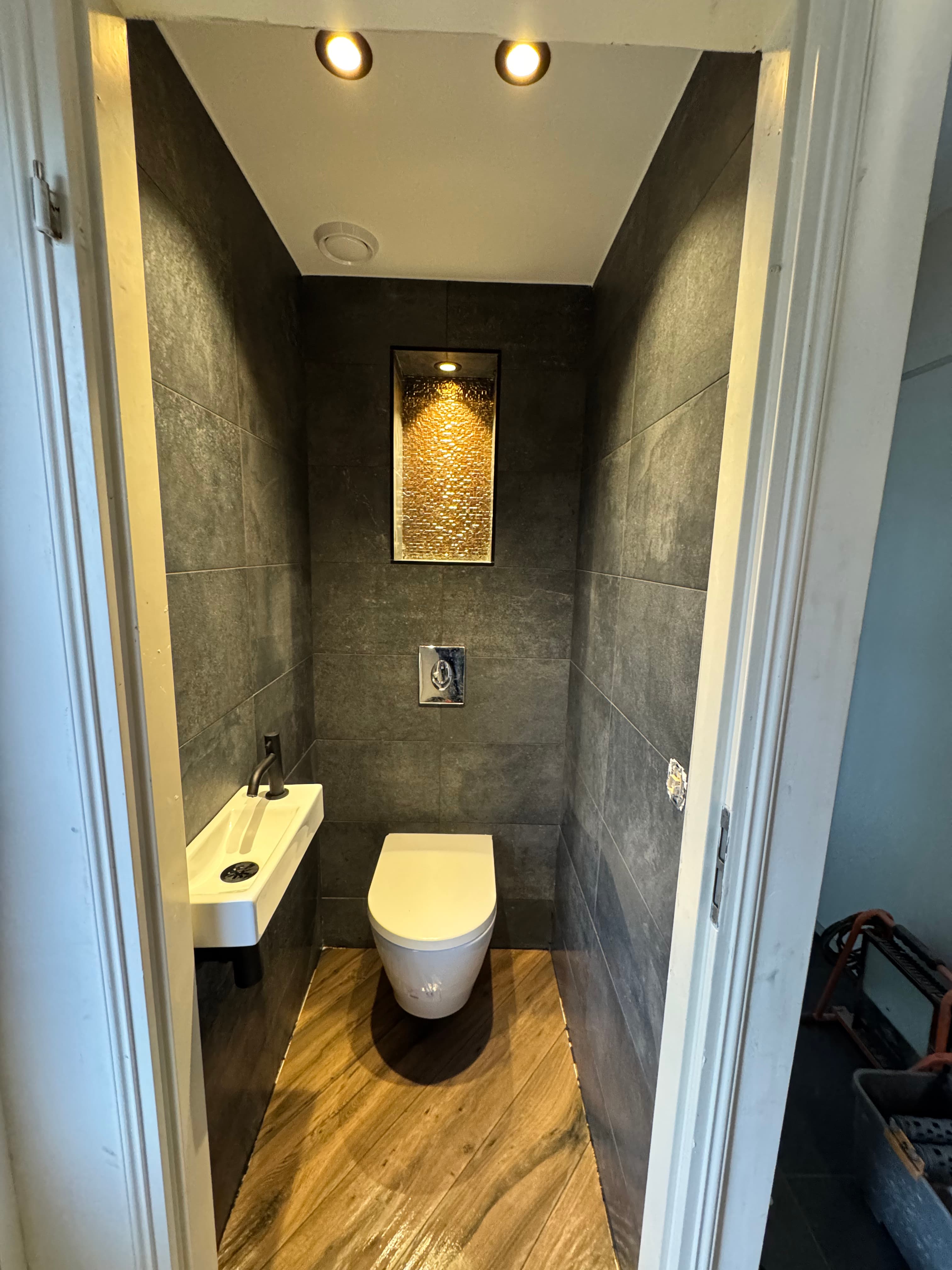 Moderne toiletruimte met donkergrijze wandtegels, houtlook vloer, wit toilet en een verlichte gouden nis.